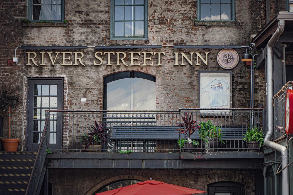 Rustic facade of River Street Inn with balcony, benches, and plants, showcasing urban charm.
