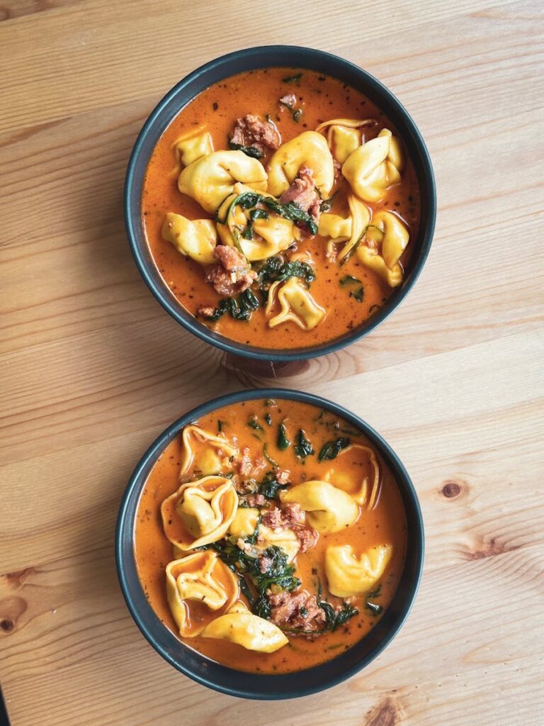 Two bowls of savory tortellini soup with sausage and herbs on a wooden surface.